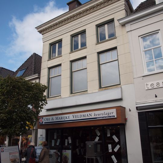 Pand met hoge voorbouw, gepleisterde gevel en zware kroonlijst onder dwars schilddak met forse hoekschoorstenen