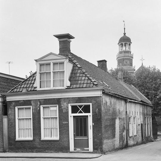 Eenvoudig pandje onder voor afgeschuind zadeldak met grote dakkapel en topschoorsteen waarop bord