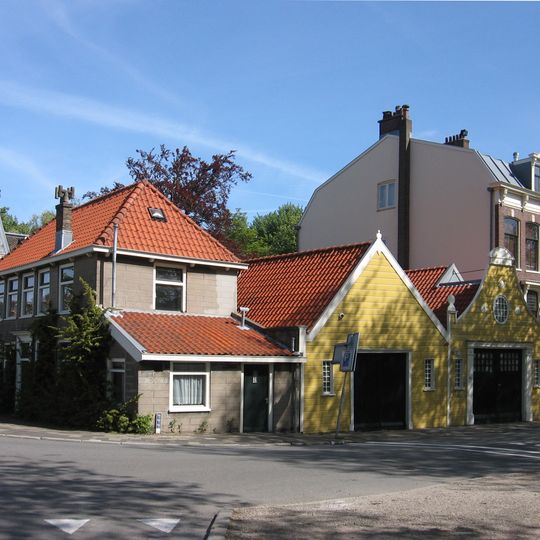 Langgerekte houten schuur onder zadeldak. Achter tuitgevel met hoekversieringen en gesneden rozetten. Met inrijpoort