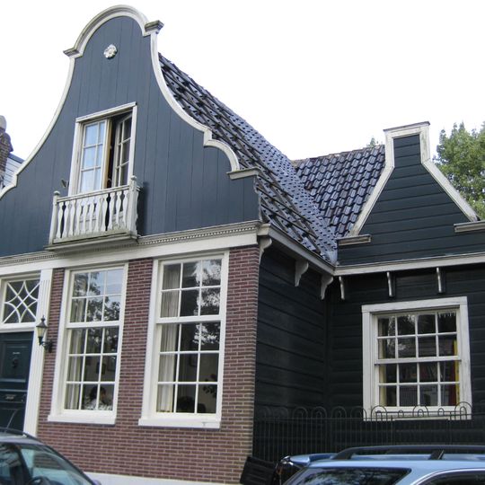 Houten dijkhuis met klokgevel en iets naar achter een tuitgevel, bovenlicht en gegroefde omlijsting, allen uit 19de eeuw, bakstenen onderpui