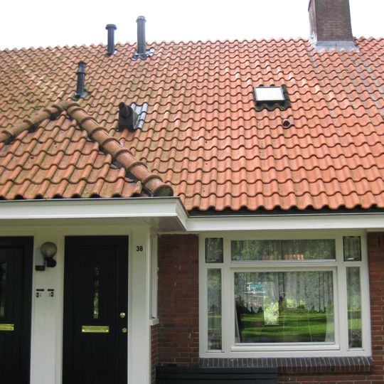 Blok met 8 woningen, gecombineerde toegangen van twee woningen onder een ingestoken schilddak, zijkant met houten topgevel, aan zuidkant toegangspoort tot binnenterrein.