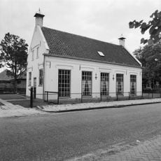 Gepleisterd dwarshuis onder zadeldak tussen puntgevels met hoekschoorstenen, waarop windkappen