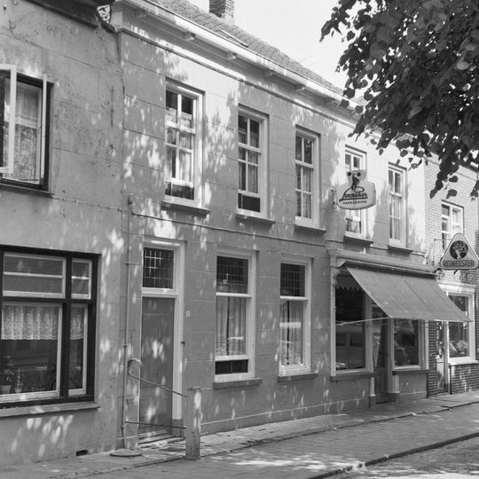 Pand met rechte bepleisterde gevel aan de straat en schilddak. Rechts winkelpui, links woonhuis. Hardstenen stoeppaal met 18e-eeuws ijzer