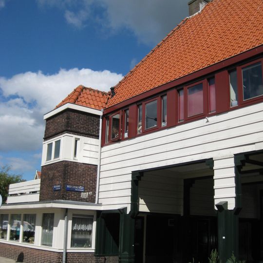 Poortgebouw met twee winkels met bovenwoningen, met veel hout zoals de zware kolommen in poort, bouwstijl Amsterdamse School, ligging aan Purmerweg centraal deel van Tuindorp Nieuwendam